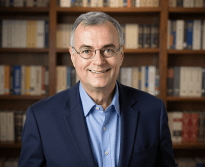 Headshot of attorney Robert P. Bergman in a navy jacket and blue shirt, standing in front of a wall of law books at his San Jose office on Ross Avenue.