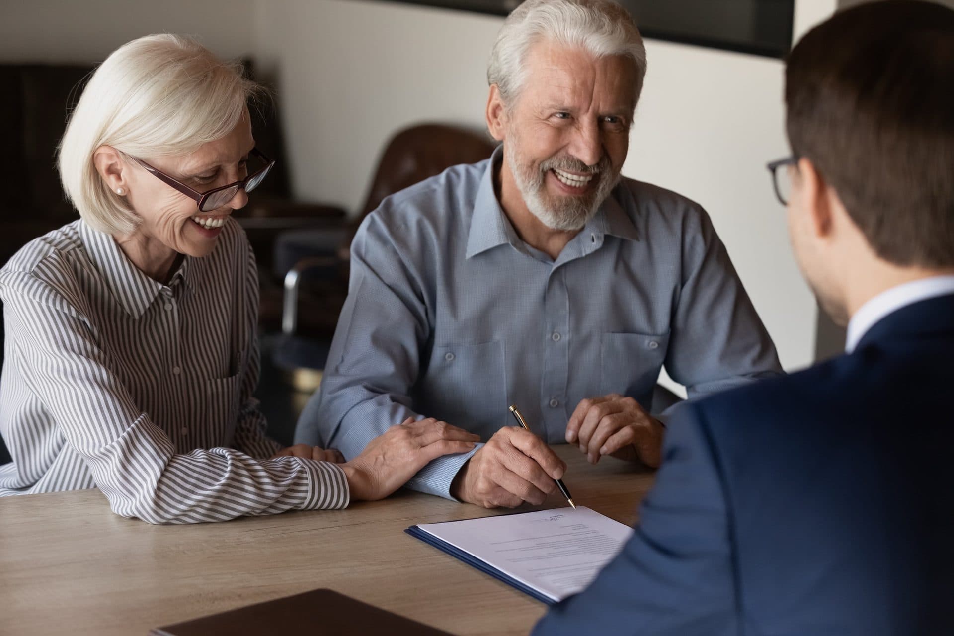 An older couple sits across a wooden conference table from an attorney in a navy suit; the attorney points at a section of an irrevocable trust they are considering modifying.