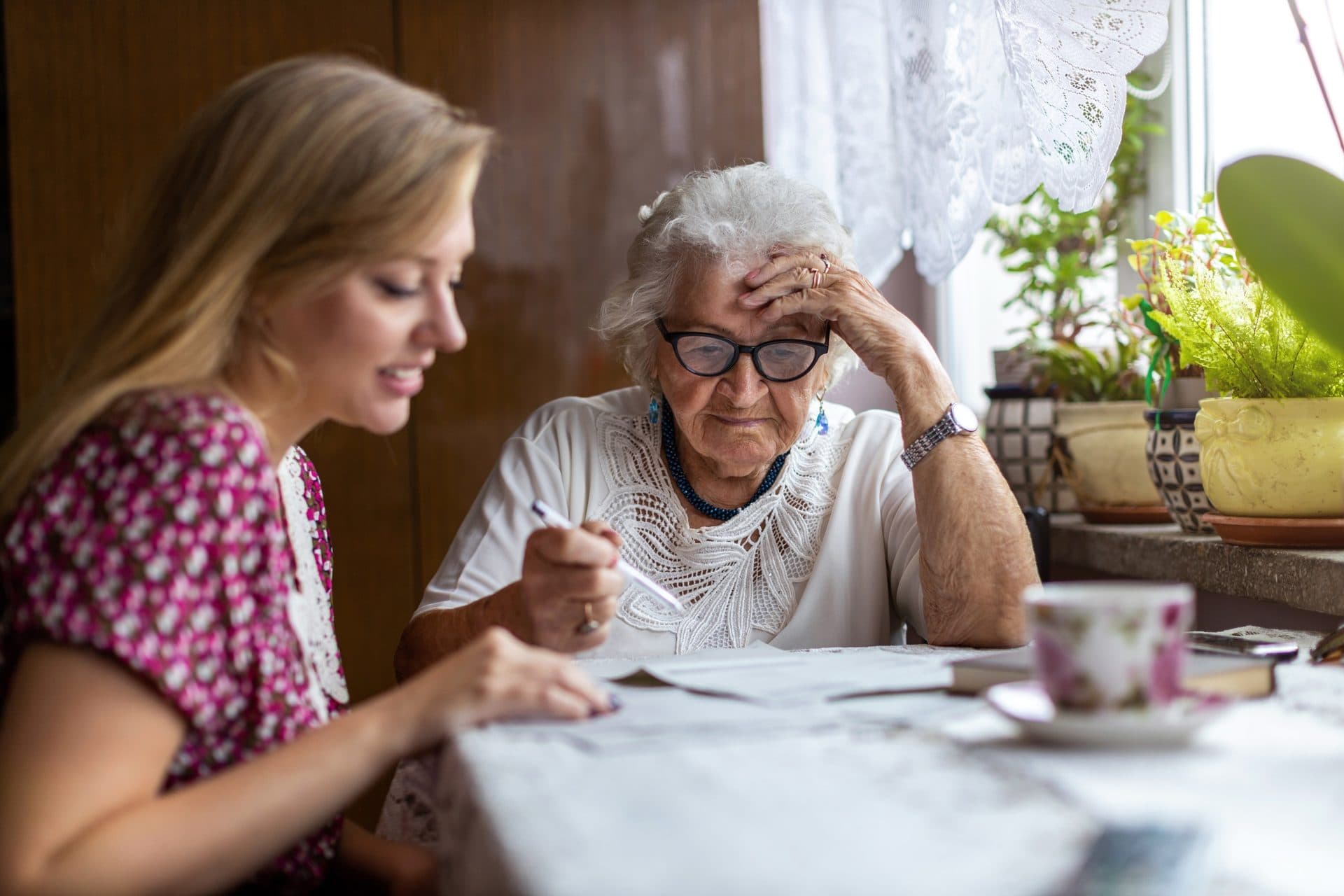 A younger blonde woman sits beside an older woman with white hair and dark glasses; both are reviewing estate planning paperwork at a sunlit table, with green plants visible behind them.